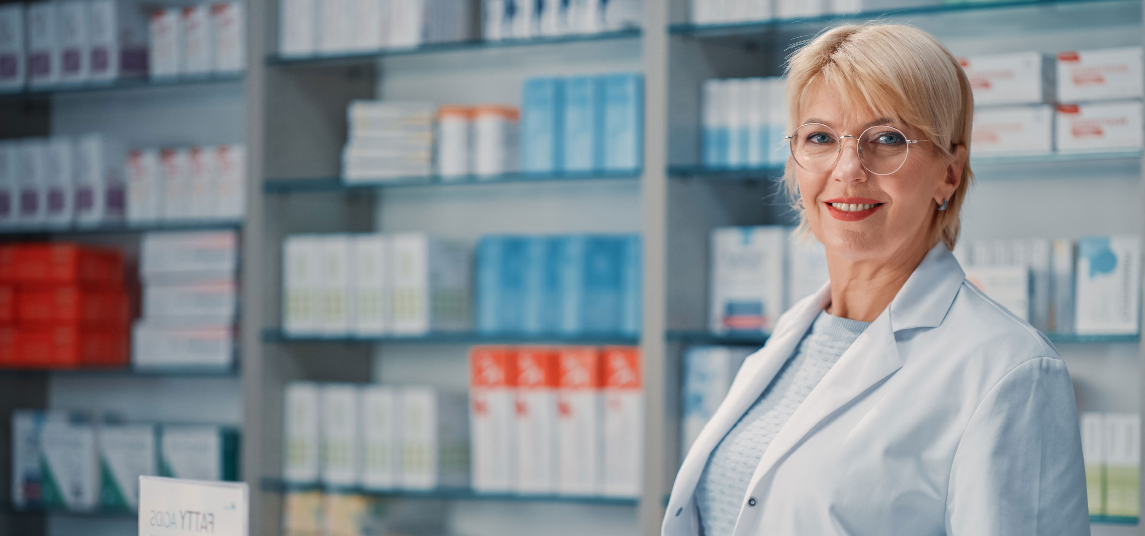 Eine Apothekerin steht in der Apotheke und lächelt in die Kamera. Hinter ihr ist eine Auswahl an Medikamenten, die nicht stark fokussiert ist.