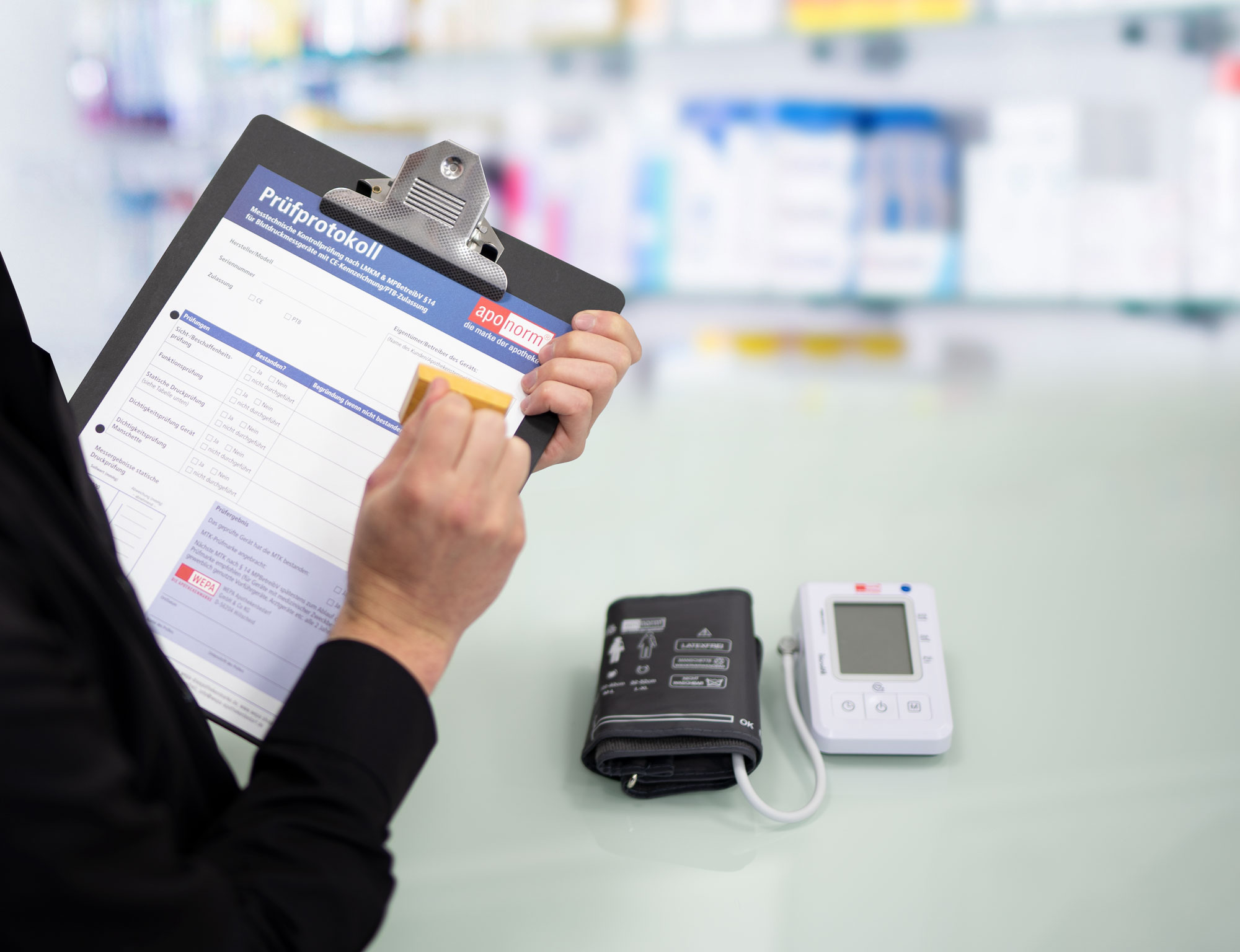 Eine Frau mit einem Klemmbrett hält gerade einen Stempel in der Hand, um ein Blatt zu stempeln. Im Vordergrund ist ein Blutdruckmessgerät zu sehen, und im Hintergrund erkennt man unscharf eine Apotheke