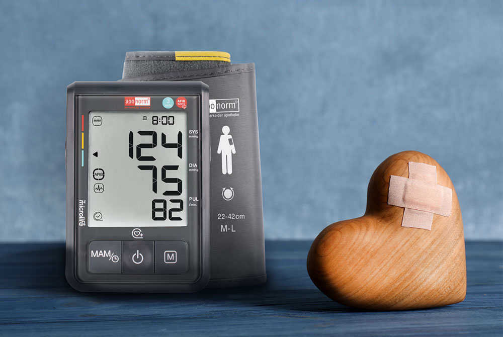 Ein Blutdruckmessgerät neben einem Holzherz, das mit zwei Pflastern als Kreuz versehen ist. Der Hintergrund ist blau