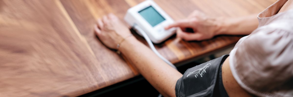 Eine Person sitzt am Tisch und hat die Manschette des Blutdruckmessgeräts um den Oberarm gelegt, während sie das Gerät drückt.