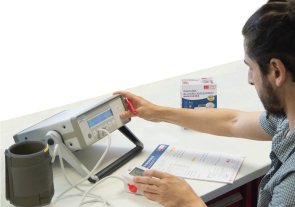 Ein Mann, der ein Gerät benutzt, das mit einem Blutdruckmessgerät und einem Kabel angeschlossen ist. Vor ihm ein Zettel und neben ihm die Packung für das Blutdruckmessgerät.