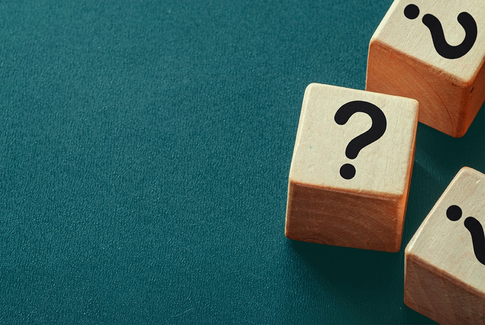 Drei Holzwürfel mit einem schwarzen Fragezeichen liegen auf einer grünen Unterlage.