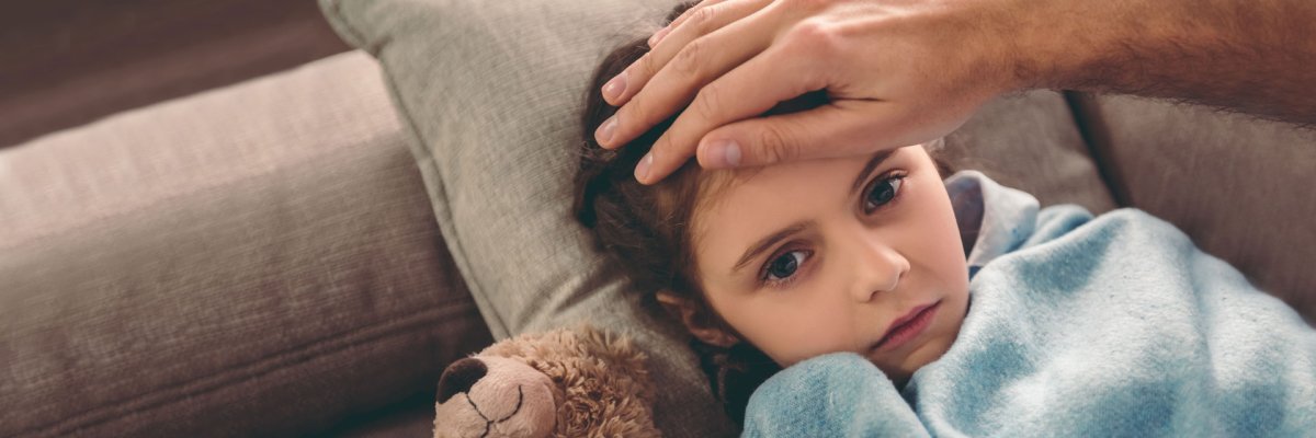 Ein Kind, das auf dem Sofa liegt, in einer blauen Decke, mit einer Hand an ihrem Kopf, misst Fieber. Sie sieht krank aus.