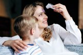 Eine Frau sitzt mit einem kleinen Jungen und misst sich selbst mit einem Fieberthermometer an der Stirn.