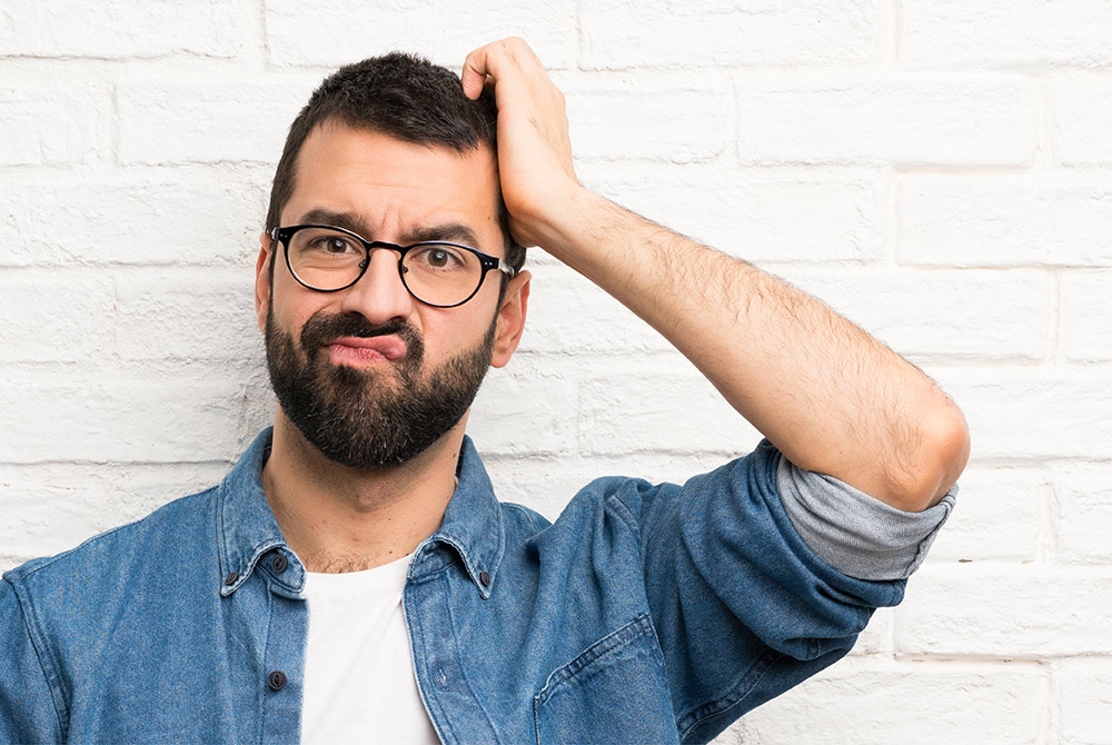 Ein Mann im weißen T-Shirt und blauen Hemd, der nachdenklich mit einer Hand an den Kopf fasst. Er steht vor einer weißen Wand und trägt eine Brille.