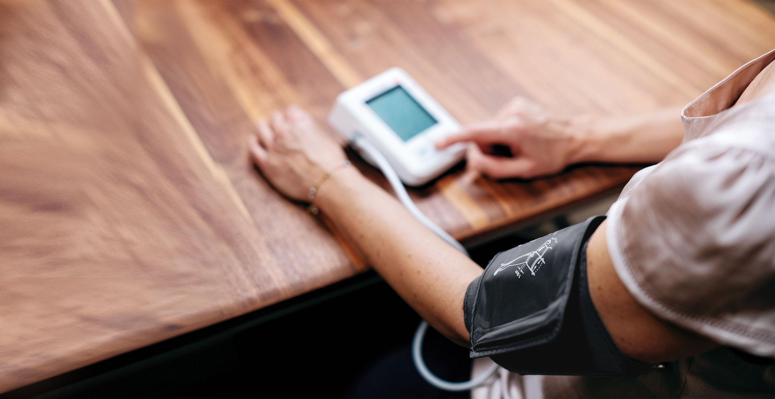 Blutdruckmessung Eine Person sitzt am Tisch und hat die Manschette des Blutdruckmessgeräts um den Oberarm gelegt, während sie das Gerät drückt.